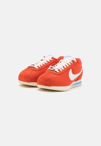 Red suede and canvas sneakers with a white swoosh logo, flat white laces, and blue accents on the heel and tongue. Classic runner design.