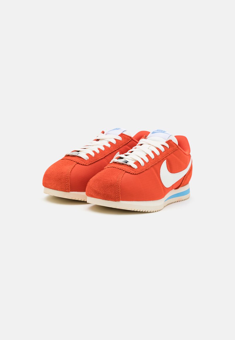 Zapatillas rojas de gamuza y lona con un logo de swoosh blanco, cordones planos blancos y acentos azules en el talón y la lengüeta. Diseño clásico de corredor.