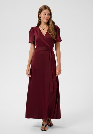 Jeune femme portant une robe portefeuille bordeaux avec des manches bouffantes et des sandales noires, debout devant un fond gris clair uni.