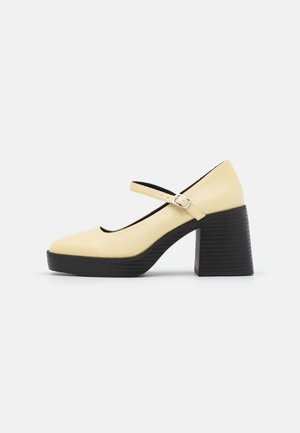 Beige Mary Jane-sko med rund tå, justerbar strop med spænde og chunky sort blokhæl og platformsål.