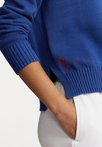 Bras dans la poche, portant un pull en maille bleu avec un logo Polo rouge et un pantalon blanc sur un fond uni.