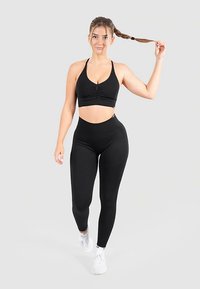 Zwarte sportbeha met geplooide details en brede bandjes, gecombineerd met zwarte leggings met hoge taille. Soepele textuur, nauwsluitend ontwerp en naadloze uitstraling.