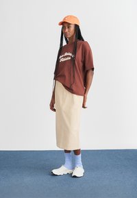 T-shirt gráfico castanho, saia midi beje, boné laranja e sapatilhas brancas com solas pretas. Meias de crews azuis visíveis. Look simples e casual.