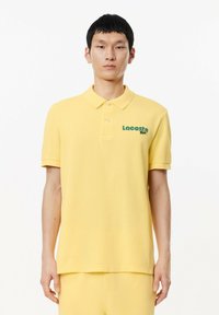Polo de color amarillo hecho de algodón con cuello, que presenta un logo verde de Lacoste en el lado izquierdo del pecho y mangas cortas.