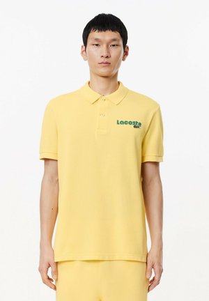 Polo de color amarillo hecho de algodón con cuello, que presenta un logo verde de Lacoste en el lado izquierdo del pecho y mangas cortas.
