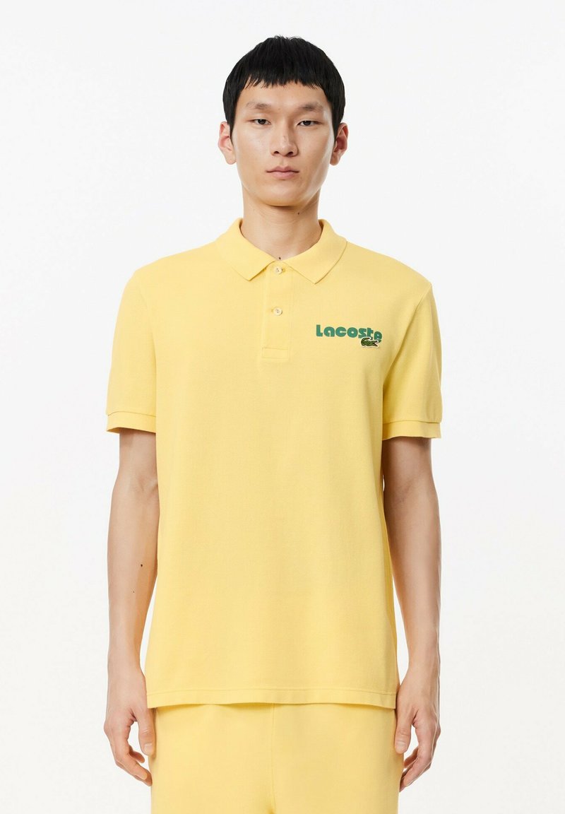 Polo de color amarillo hecho de algodón con cuello, que presenta un logo verde de Lacoste en el lado izquierdo del pecho y mangas cortas.