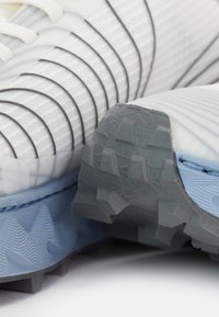 Weiße Sportschuhe mit strukturiertem Obermaterial, schwarzen Linienmustern, hellblauer Sohle und robustem grauen Außensohle. Fokus auf Details des Grip-Designs.