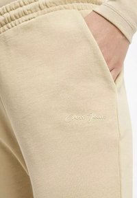 Beżowe spodnie dresowe o miękkiej teksturze, z elastycznym paskiem w talii i boczną kieszenią. Logo "Cross Jeans" wyhaftowane na białym tle na nodze.
