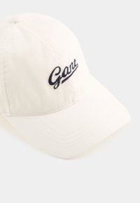 Hvit bomull baseball cap med buet skygge. Har brodert "Gant"-logo i marineblått. Sømdetaljer på skyggen gir tekstur.