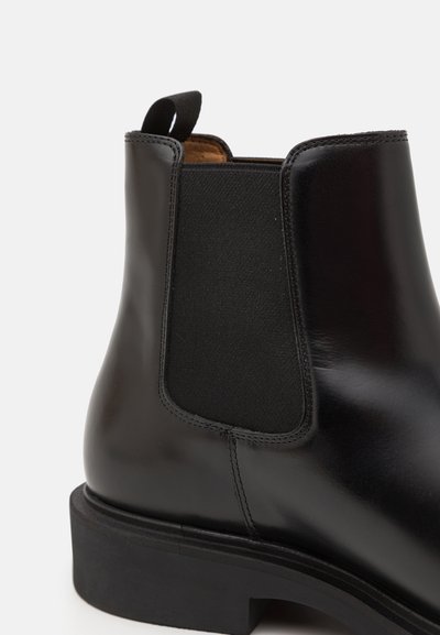 Botín de cuero negro hasta el tobillo con paneles laterales elásticos, puntera redondeada y un tacón bajo apilado. Textura suave con una etiqueta de tiro para fácil colocación.