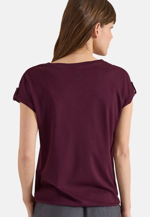 Mujer con cabello largo y castaño que lleva una camisa de manga corta morado oscuro con pestañas abotonadas en las mangas, vista desde atrás sobre un fondo liso.