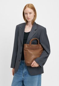 Brązowa skórzana torba tote z zaokrąglonymi rączkami, miękką teksturą i minimalistycznym designem. Nosi się ją z szarym żakietem w prążki i czarną bluzką.