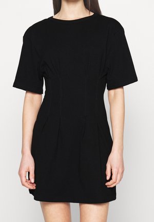 Vrouw draagt een korte, zwarte mini-jurk met een aangesloten taille en subtiele stiksel details, staand tegen een effen lichte achtergrond.
