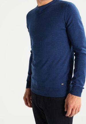 Marineblå crewneck sweater i blød strik, med ribkant ved kanten og manchetterne, samt et lille logo mærke nederst til venstre.