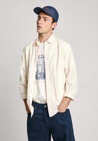 Camisa crema de botones sobre una camiseta blanca con gráfico y pantalones azul marino, complementada con una gorra de mezclilla oscura con bordado naranja.