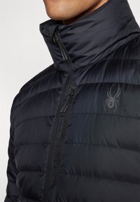 Dunkelblaue Steppjacke mit hohem Kragen, ausgestattet mit Reißverschlüssen und einem dezenten Spinnenlogo auf der Brust. Weicher, gesteppten Textur.