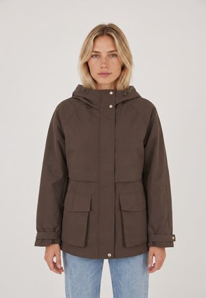 Donna bionda che indossa una giacca marrone con cappuccio e grandi tasche a patta e jeans azzurri chiari, in piedi davanti a uno sfondo uniforme.