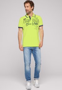 Limegroene polo met marineblauwe accenten en gedrukte tekst, gecombineerd met lichtblauwe jeans. Het shirt heeft een klassieke kraag en korte mouwen.