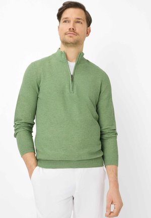 Mand iført en lysegrøn kvart-lynlåstrøje over en hvid skjorte med hvide bukser, stående med hænderne i lommerne og ser let opad.