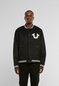 Must maik Varsity jakk, millel on valge triibuline ribikrae ja mansetid. Esiküljel on silmapaistev logo ja kaks eesmist taskut.
