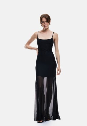 Frau trägt ein schwarzes ärmelloses Kleid mit transparentem Mesh-Rocküberzug und steht mit einer Hand auf der Hüfte vor einem schlichten Hintergrund.