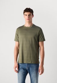 Moški nosi olivno zeleni kratkosleeve majico z teksturo in modre jeans hlače, stoji ob preprosti svetli ozadju.