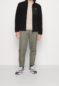 Chaqueta de forro polar negra con un bolsillo en el pecho con cremallera, combinada con una camisa de color beige claro y pantalones verde oliva. El calzado presenta un diseño en blanco y negro.