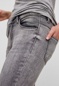 Jeans de ganga cinza com um corte slim. As características incluem um design padrão de cinco bolsos, textura desgastada e detalhes em metal prateado.