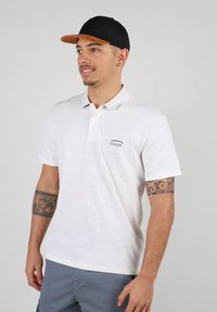 Polo blanc à manches courtes, avec un petit logo Oxbow. Casquette noire à visière plate avec des accents marron. Peau bronzée, tatouage visible.