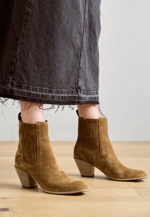Bottes chelsea en daim marron avec un bout pointu et un talon épais en bois. La tige présente des coutures verticales et une texture lisse.