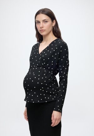 Gravid kvinde iført en sort langærmet top med hvide prikker og et sort nederdel, stående mod en ensfarvet hvid baggrund.