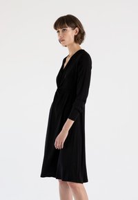 Abito nero realizzato in tessuto morbido, con scollatura a V, maniche lunghe e vita arricciata. Gonna svasata che arriva sopra il ginocchio. Design semplice.