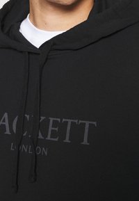 Černá mikina s kapucí a šňůrkami, s velkým šedým potištěným logem "HACKETT LONDON" na přední straně, vyrobená z měkké látky.