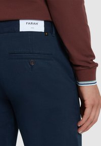 Pantaloni blu navy con una trama liscia. Presentano una tasca sul retro con un bottone e un'etichetta bianca con il marchio "FARAH" in vita.