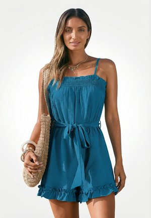 Kobieta ubrana w bezrękawnikowy turkusowy kombinezon z paskiem w talii i falbaną na dole, niosąca plecioną torbę i nosząca bransoletki oraz naszyjnik.