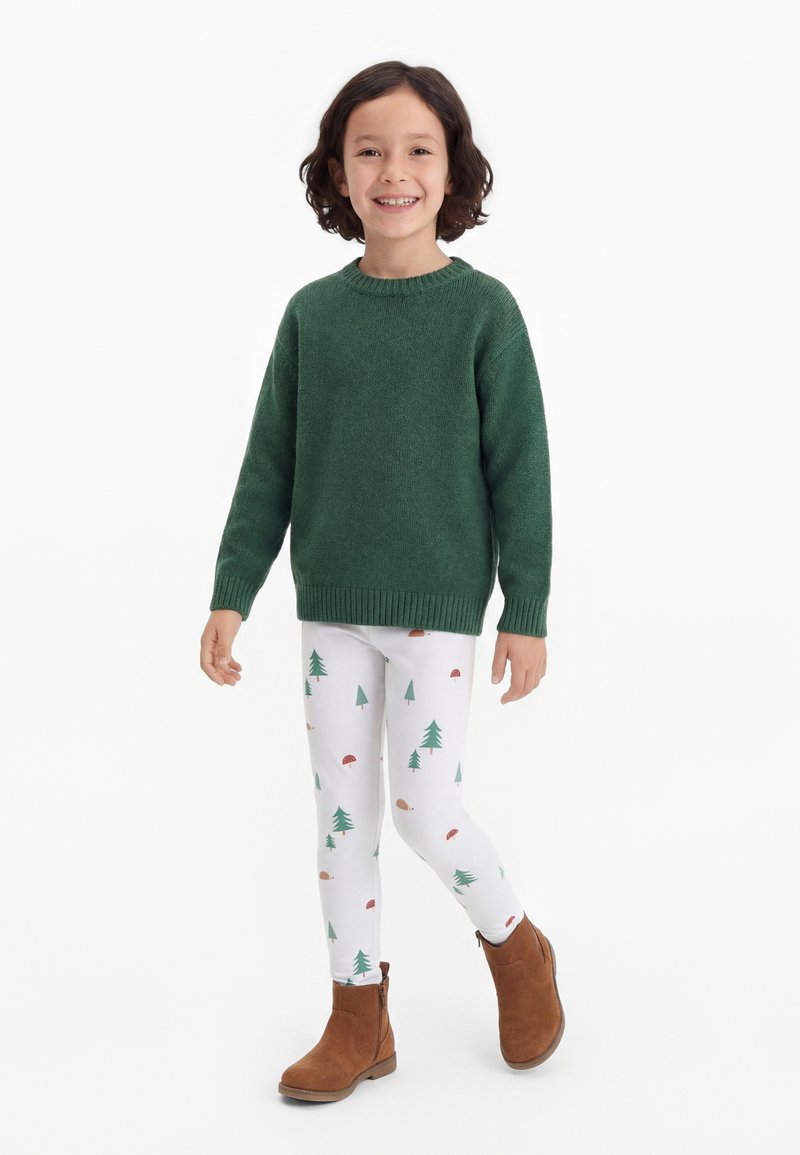 Enfant souriant aux cheveux foncés portant un pull vert, des leggings blancs avec des motifs d’arbres et de champignons, et des bottines marron, debout sur fond blanc.
