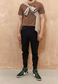 T-shirt marrone con logo nero, pantaloni jogger neri con vita elasticizzata e tasche, e scarpe alte verde/nero con dettagli bianchi.