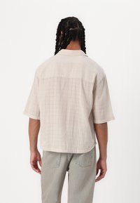 Beige Kurzarmhemd mit einem strukturierten Rippmuster, lockerer Passform und spitzem Kragen, kombiniert mit hellblauen Jeans.