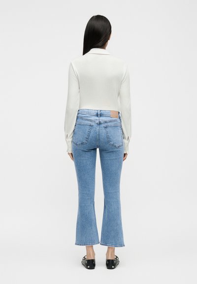 Γυναίκα με μακριά σκούρα μαλλιά, ντυμένη με λευκό μακρυμάνικο τοπ, ανοιχτόχρωμα γαλάζια παντελόνια καμπάνα έως τον αστράγαλο και μαύρα επίπεδα παπούτσια, στέκεται με την πλάτη προς τα εμπρός.