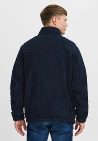 Veste en polaire bleu marine avec un col haut, surface texturée et poignets élastiques. Elle présente une coupe décontractée et un design simple et uni.