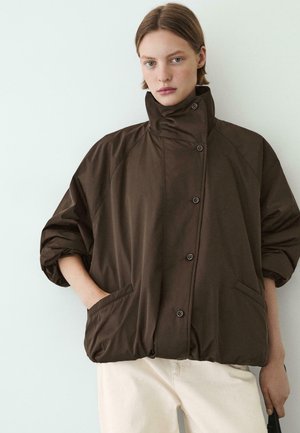 Jeune personne aux cheveux courts bruns portant une veste matelassée boutonnée marron foncé avec un col montant et un pantalon blanc, debout devant un fond clair.