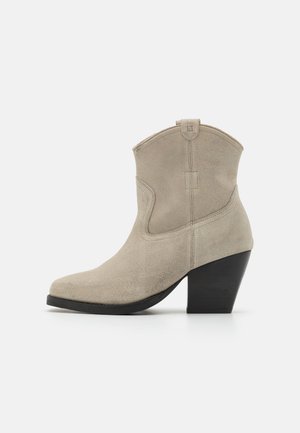 Bottine en daim beige avec un bout pointu, des côtés incurvés et un talon noir texturé. Comprend des coutures et une boucle à l'arrière.