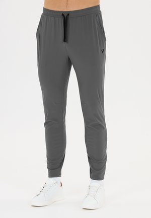 Homme portant un pantalon de jogging slim gris foncé avec un cordon noir et des baskets blanches sur un fond blanc uni.