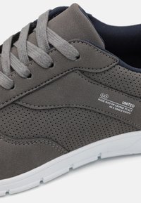 Grey sneaker with perforated texture, fabric upper, and white rubber sole. Features grey laces and branding tag on the side.