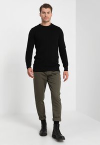 Zwarte trui met een gestructureerd zigzagpatroon, nauwsluitende snit; olijfgroene broek, tapering ontwerp; zwarte combat boots; effen achtergrond.