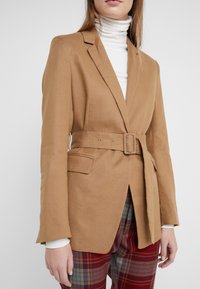 Brązowa marynarka z paskiem, o dopasowanym kroju, wykonana z teksturowanej tkaniny. Posiada klapy typu notch oraz dwie przednie kieszenie z widocznym przeszyciem.