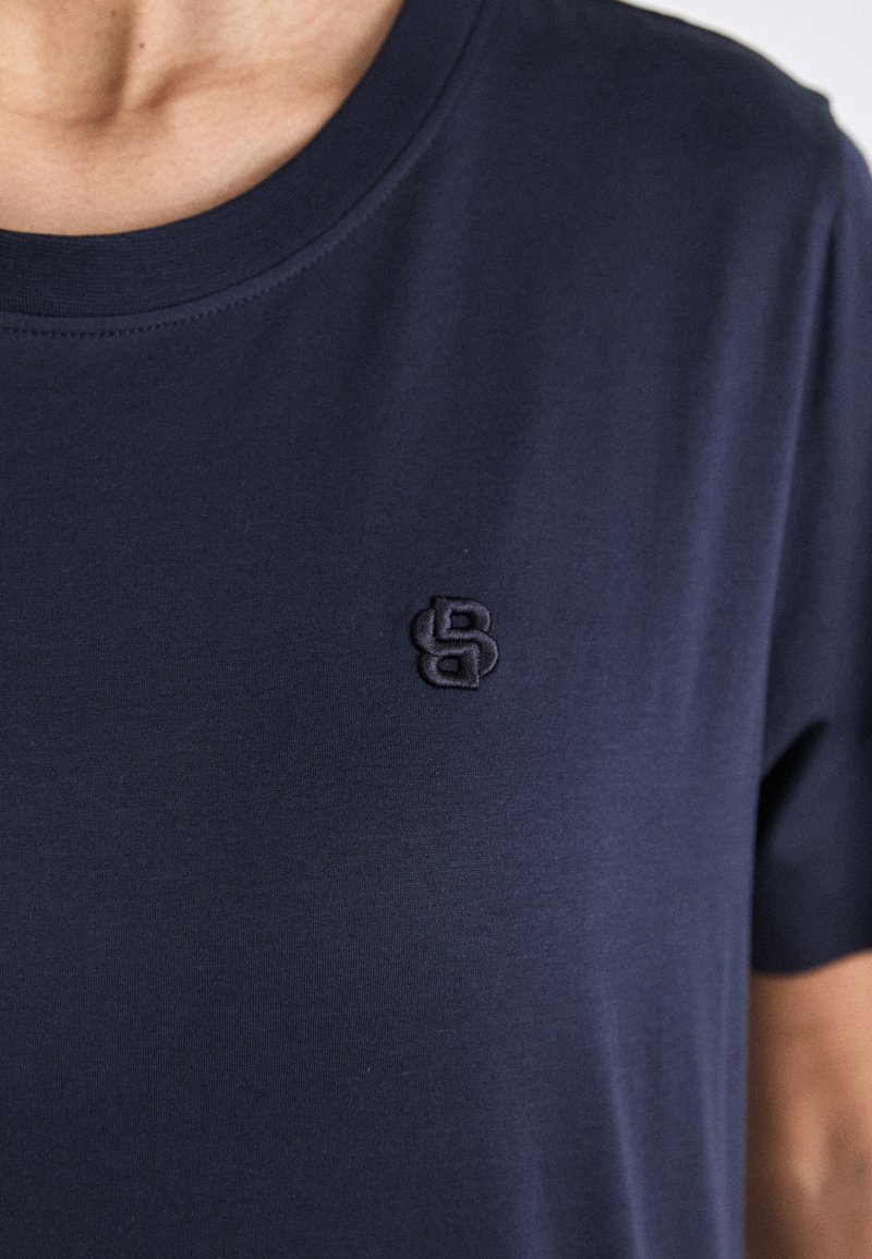 Tiefblaues Baumwoll-T-Shirt mit Rundhalsausschnitt und einem gestickten Logo in Form von ineinander verschlungenen Buchstaben auf der Brust.
