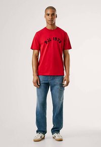 Raudona medvilninė marškinėliai su apvalia iškirpte ir juodu užrašu „PJL 1973“, derinama su mėlynais džinsais iš džinso audinio, turinčiais priekinės kišenes ir laisvesnį kirpimą.