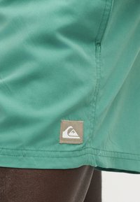 Σορτς από τιρκουάζ πράσινο ύφασμα με μικρό τετράγωνο μπεζ λογότυπο κοντά στο τελείωμα πάνω από σκούρο δέρμα χρώματος δέρματος.