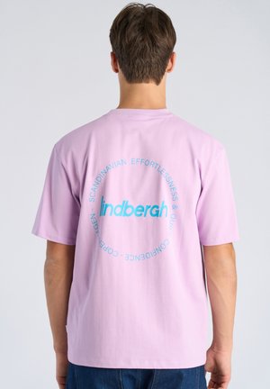 Uomo che indossa una maglietta viola chiaro con testo circolare blu sul retro con la scritta "indbergh" e "Scandinavian effortlessness & quiet confidence Copenhagen".